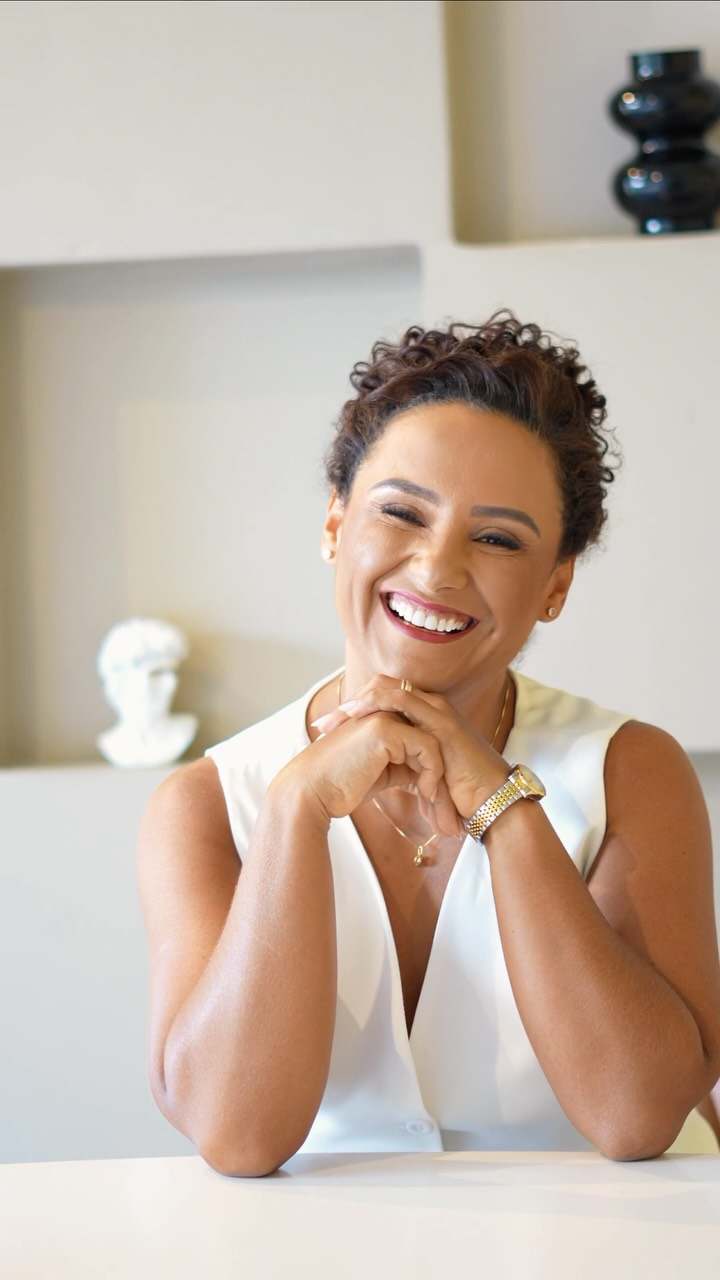 Retrato profissional de Jacque Oliveira sorrindo, com blusa branca e as mãos próximas ao rosto.
